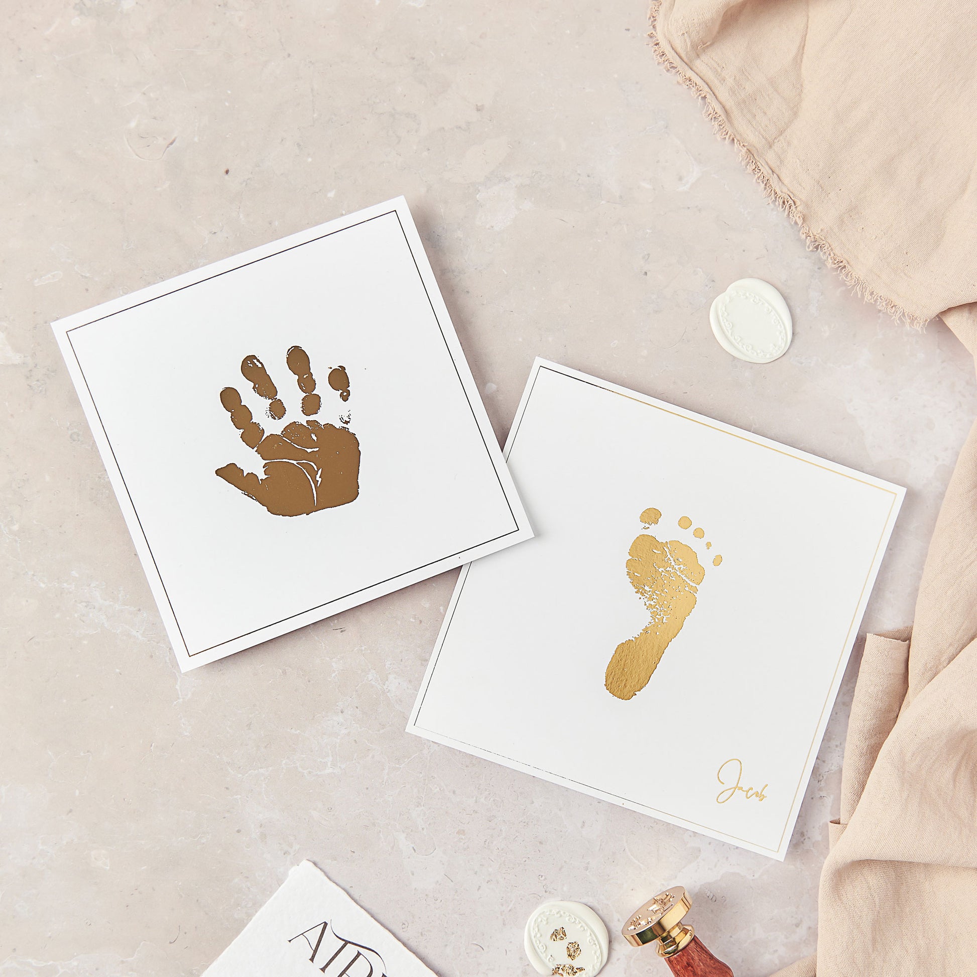 This photograph shows 2 personalised keepsakes, both at 5 inches square. To the left of the photograph is a foil hand print keepsake, created in gold foil on to white card stock. To the right is a seperate foil keepsake this time showing a gold foiled baby footprint on white cardstock. The foil footprint is fused centrally within the 5 inch square white cardstock. Within the bottom right corner of this print it is further personalised with the baby's name "Jacob" in a gold foil script font.