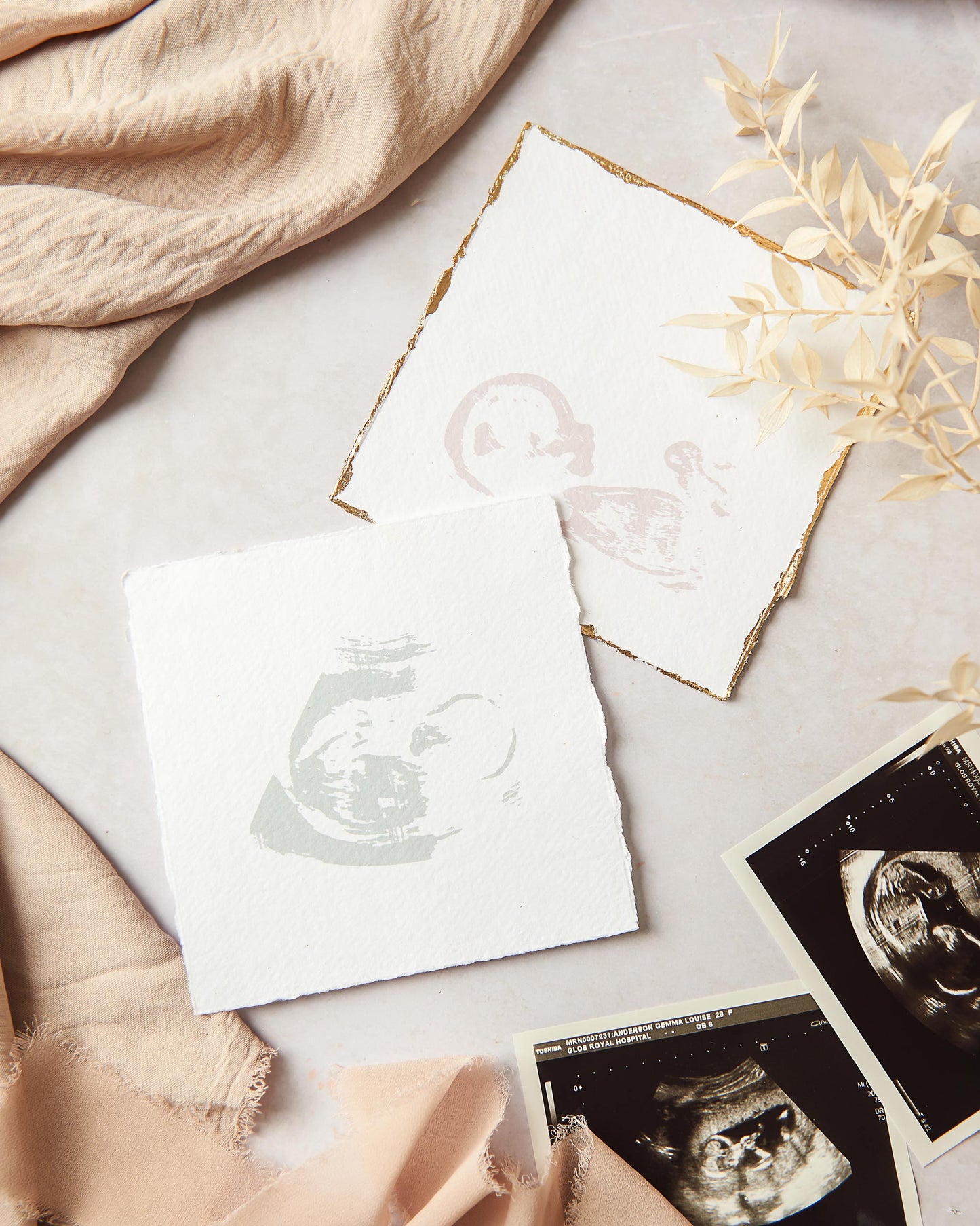 A photograph showing 2 baby scan keepsake examples. To the foreground the keepsake is shown with the baby scan replicated in light sage green on white handmade paper with it's natural feathery deckle edges. Behind this you can see the other keepsake example, the baby scan is replicated in a light dusky pink ink on to white handmade paper with it's deckle edges gilt in a gold colour metal leaf. Surrounding the keepsakes are 2 original baby scans for reference points and fabric and dried flower stem props.