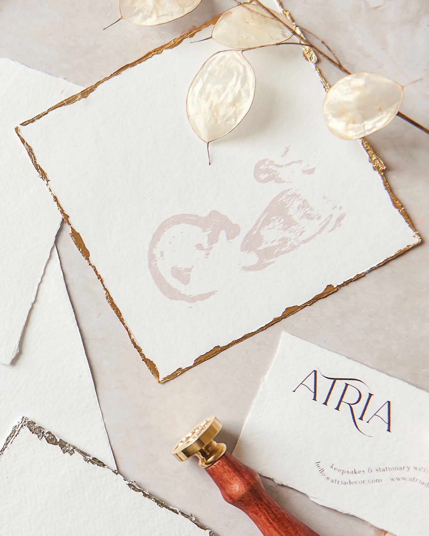 A close up photograph of a baby scan keepsake on white handmade paper, with gold leaf gilt deckle edges and the silhouette of the baby printed in a light dusky pink ink. Other handmade papers, a brass wax seal stamp and a dried stem of lunaria honesty surround the sentimental keepsake.