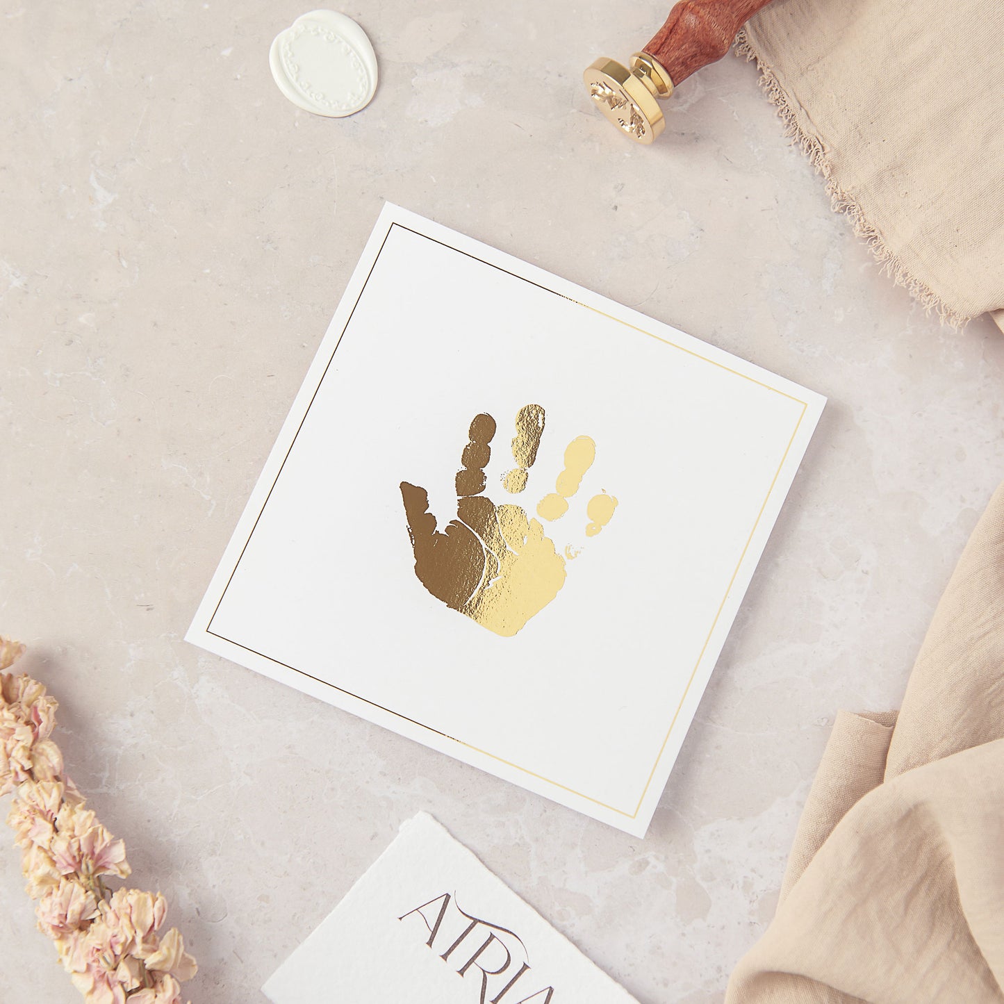 A flat lay photograph showing our 5 inch square personalised hand print keepsake. The Hand print keepsake is shown in Gold and white, where the baby's handprint is recreated to scale in metallic gold foil which is fused on to smooth white cardstock centrally. The personalised hand print keepsake is unframed and is shown resting on a light pink marble style back drop. Fabric, wax stamp seals, a floral stem and Atria UK branded card surrounds the keepsake to decorate the edges of the photograph.
