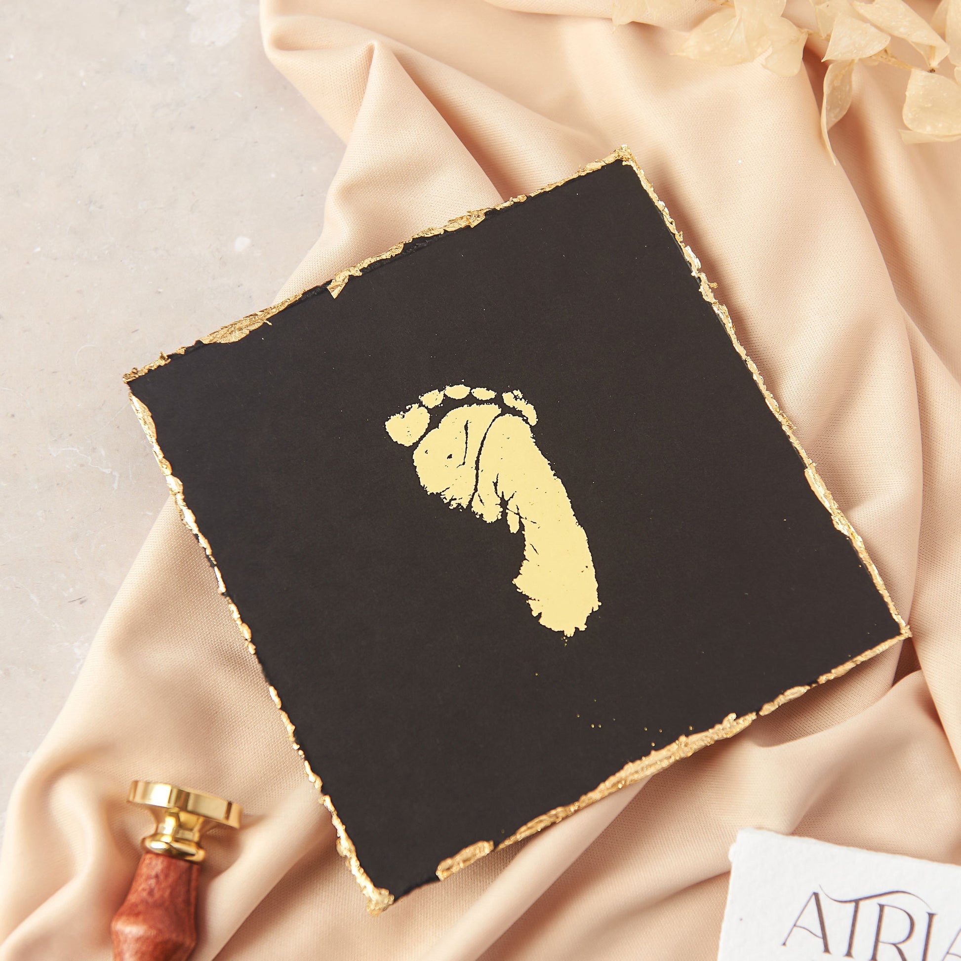 This photograph shows a baby's footprint which has been recreated in gold foil on to black cardstock. The paper keepsake is 5 inches square and has been finished with a hand deckled gold gilded edge which is an add on option to a standard foil print keepsake. The keepsake is shown on a draped neutral fabric backdrop.