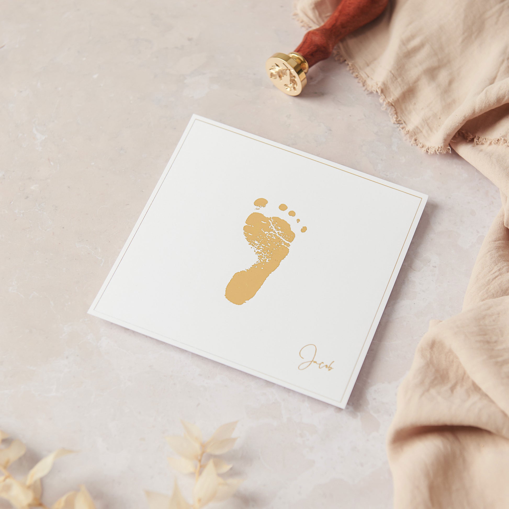 A flat lay photograph showing our 5 inch square personalised foot print keepsake. The foot print keepsake is shown in Gold and white, where the baby's footprint is recreated to scale in metallic gold foil which is fused on to smooth white cardstock centrally. The foot print keepsake is further personalised with the baby's name "jacob" in a gold foiled script font to the bottom right corner of the cardstock. The keepsake is unframed and is shown resting on a light pink marble style back drop.