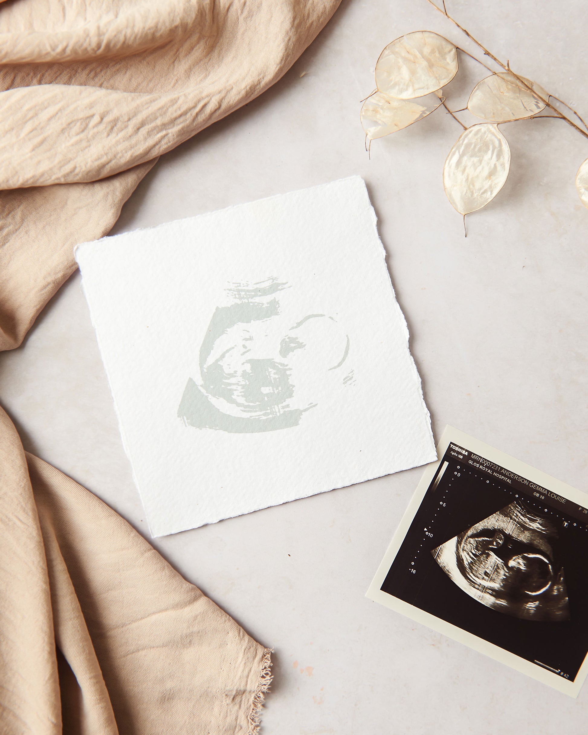 A photograph showing a handmade paper baby scan keepsake. The keepsake is appoximately 5 inches square and has the silhouette of the baby printed centrally in a light sage green ink, on to a natural handmade paper with its feathery deckle edges. Surrounding the keepsake in the photograph are the original scan to show the reference point along with fabric and a dried stem of lunaria honesty.