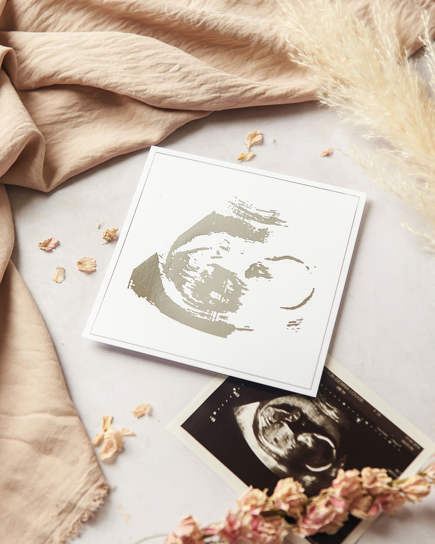 Photograph shows our foil babyscan keepsake, next to an original baby scan and on a neutral background. Our foil baby scan keepsake is shown in silver foil on white 5 inch square cardstock.