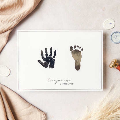 The hand and foot print pair keepsake photographed is shown in silver foil on white A5 paper size cardstock. The print photographed includes a baby's hand and foot print side by side in silver foil, positioned centrally on to A5 size white cardstock. The keepsake is further personalised in silver foil to include the baby's name "luna joan rose" in script font and date of birth "2 June 2021" in sans-serif Capital case font. This personalised text is positioned centrally at the bottom of the A5 cardstock.